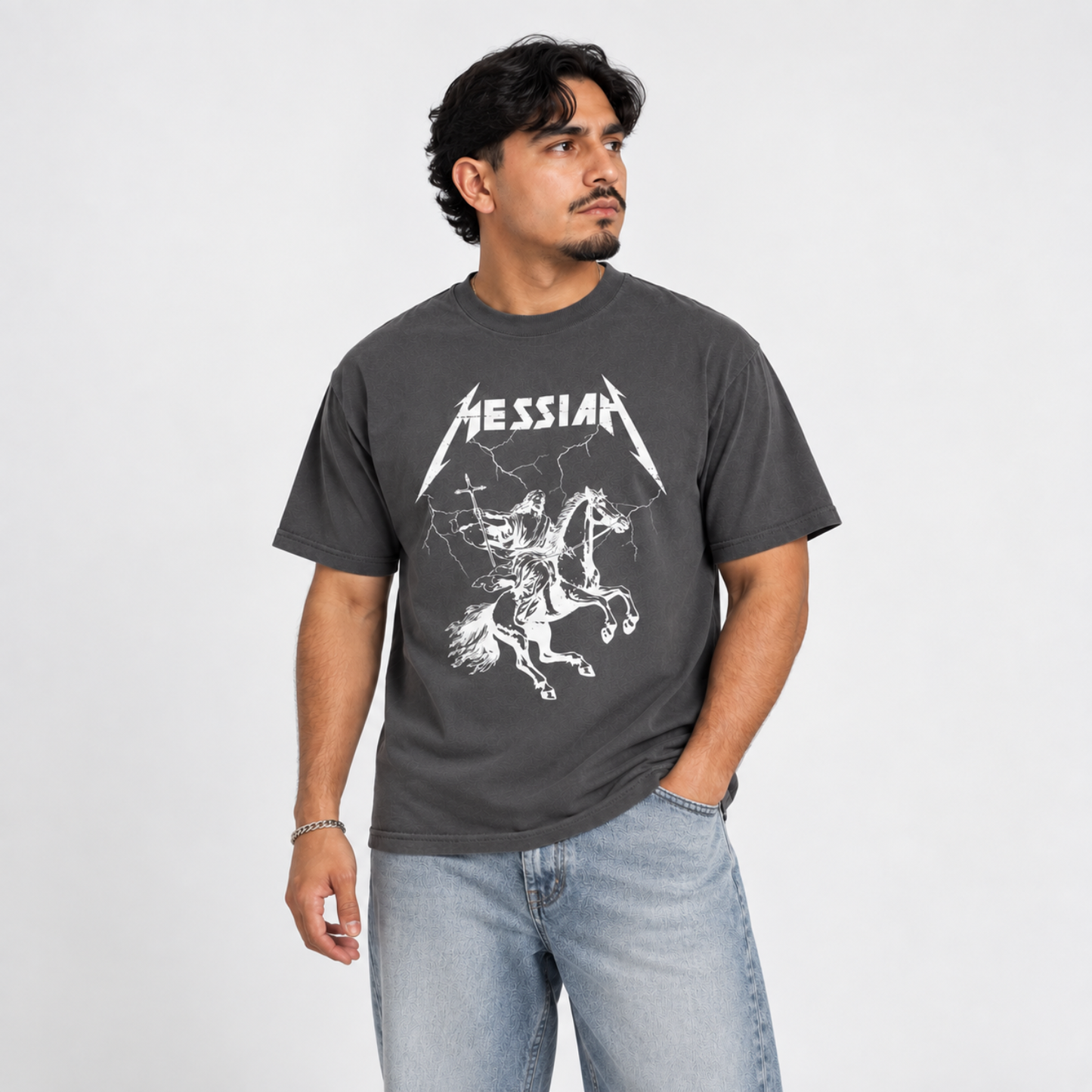 A latino man wearing a gray graphic t-shirt with a white 'Messiah' design, paired with light blue jeans, standing against a light gray studio background.